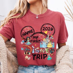 A person wearing a red t-shirt with Disney-themed graphics and text, including the words "Name my first Disney trip" and "2020".