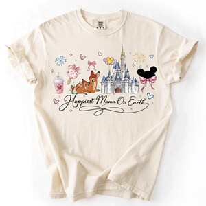 A white t-shirt with a printed design featuring Disney characters and landmarks, along with the text "Happiest Mama On Earth".