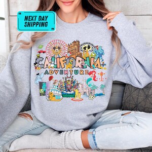 A person wearing a gray sweatshirt with a colorful graphic design depicting various California-themed elements, such as a ferris wheel, roller coaster, and palm trees.