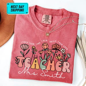 May include: A red Comfort Colors t-shirt with the words "TEACHER Mrs. Smith" in a colorful, retro font, surrounded by a floral design. The shirt also has the text "TEACH THEM. LOVE THEM. WATCH THEM GROW". A silver necklace is draped across the shirt.