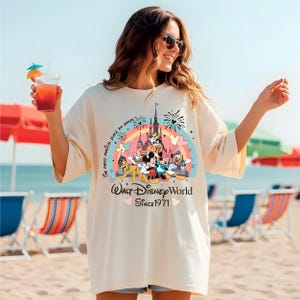 A woman wearing a white t-shirt with a Disney World logo and text, standing on a beach with colorful umbrellas and chairs in the background.