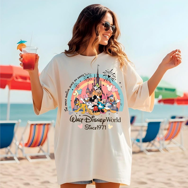 A woman wearing a white t-shirt with a Disney World logo and text, standing on a beach with colorful umbrellas and chairs in the background.