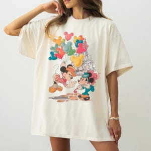A woman wearing a white t-shirt with a colorful graphic of Mickey Mouse and Minnie Mouse surrounded by balloons.