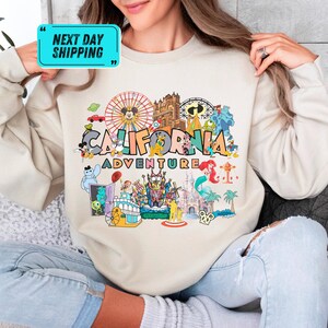 A person wearing a white sweater with a colorful graphic design depicting various Disney characters and landmarks, including a ferris wheel, castle, and palm trees, along with the text "California Adventure".