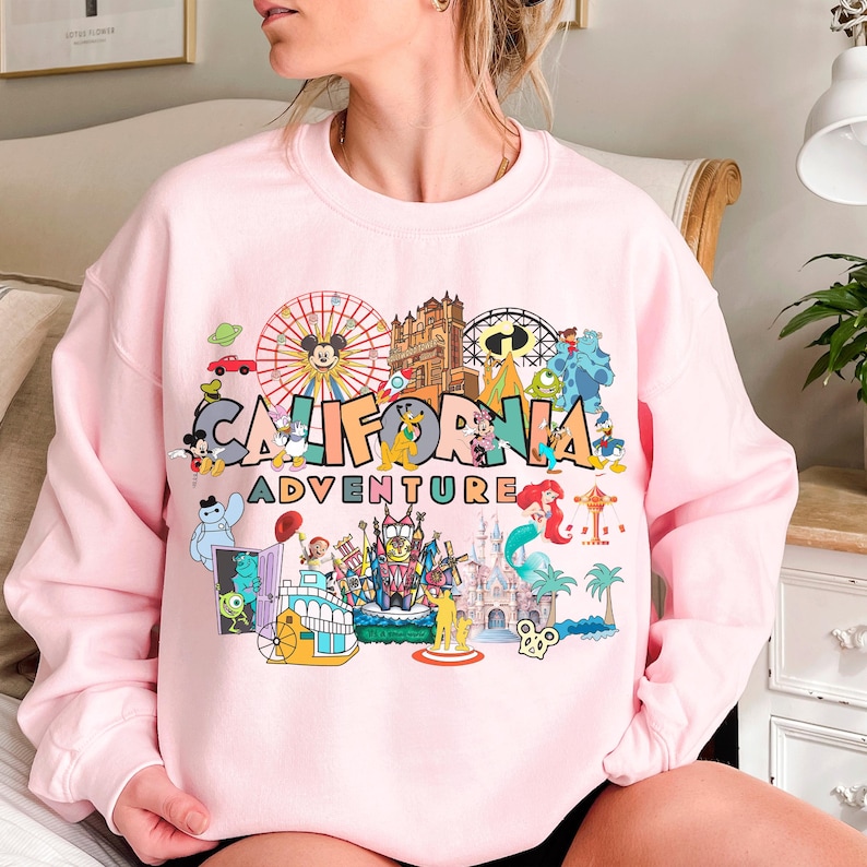 A woman wearing a pink sweatshirt with a colorful graphic design depicting various Disney and amusement park attractions, including Mickey Mouse, the California Adventure, and other iconic landmarks and characters.