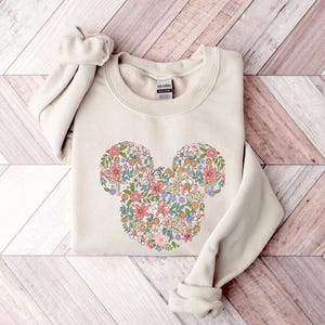 May include: A beige sweatshirt featuring a floral Mickey Mouse design. The flowers are in shades of pink, purple, yellow, and green.