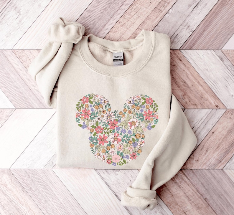 May include: A beige sweatshirt featuring a floral Mickey Mouse design. The flowers are in shades of pink, purple, yellow, and green.