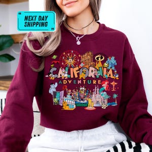 A woman wearing a red sweater with a colorful graphic design depicting various landmarks and characters from California, including the Golden Gate Bridge, Disneyland, and Mickey Mouse.
