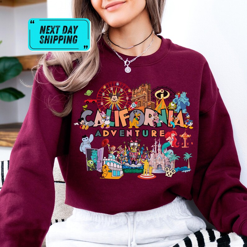 A woman wearing a red sweater with a colorful graphic design depicting various landmarks and characters from California, including the Golden Gate Bridge, Disneyland, and Mickey Mouse.