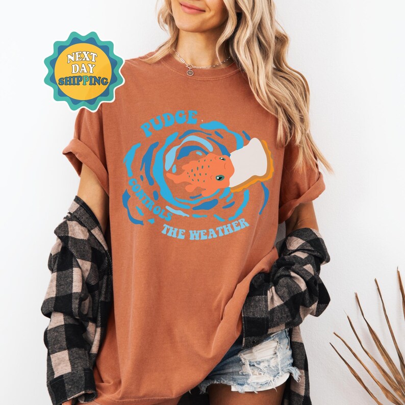 以下が含まれることがあります： さび色のTシャツに、口にパンの切れ端をくわえた魚のグラフィックがプリントされています。Tシャツの文字は「Fudge Controls The Weather」です。