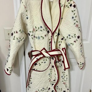 May include: Cream-colored quilted robe with floral embroidery in blue, pink, and green. The robe has a burgundy trim and a matching belt. It is hanging on a white hanger against a white door.