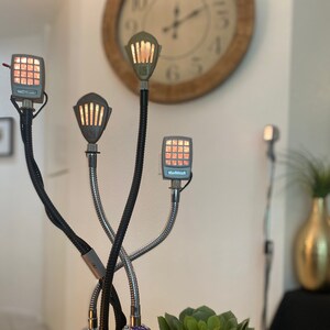 May include: A black metal desk lamp with three adjustable arms. Each arm has a silver light fixture with a grid of small, warm-toned LED lights. The lamp is on a black base with a small purple pouch and a QR code sticker.