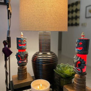 May include: Two tiki-style candle holders with red and blue accents, a white candle with three wicks, and a brown lamp with a beige lampshade on a black table.