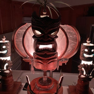 May include: Three tiki lamps with carved wooden faces and glowing lights. The centre lamp has a larger face with a circular, translucent shade. The lamps are on wooden bases.