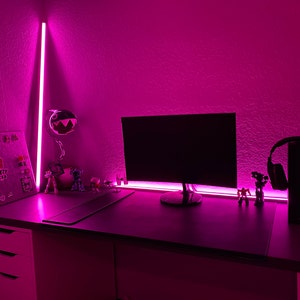 May include: A pink-lit desk with a computer monitor, a keyboard, a mouse, a headset, and a few small figurines. The desk has a white drawer unit on the left side and a black surface.
