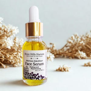 May include: A clear glass bottle of River Hills Harvest American Elderflower Face Serum. The bottle has a gold cap and a white dropper. The label includes the text "Healing and Rejuvenating" and is .5oz (14.78mL).