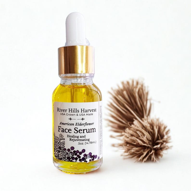 May include: A clear glass bottle of American Elderflower Face Serum with a gold cap and white dropper. The label reads "River Hills Harvest" and "Healing and Rejuvenating." The bottle contains a yellow liquid. A dried plant is in the background.