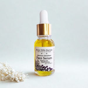 May include: A clear glass bottle of River Hills Harvest American Elderflower Face Serum. The bottle has a gold cap and white dropper. The label includes text: "Healing and Rejuvenating" and "5oz. (14.78ml)".