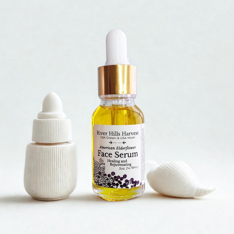 May include: A clear glass bottle of face serum with a gold cap and white dropper, next to a white ceramic bottle and a seashell. The serum bottle has a label that reads "American Elderflower Face Serum".
