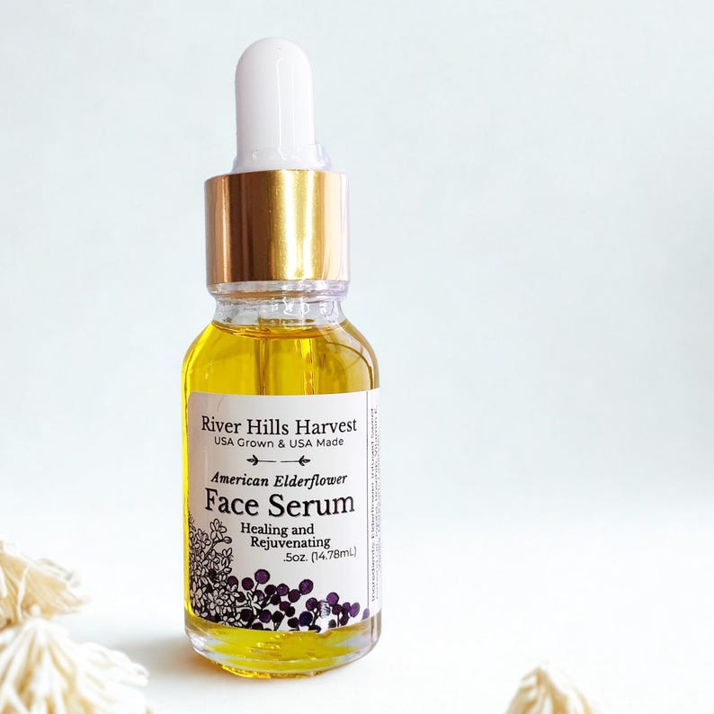 May include: A clear glass bottle of American Elderflower Face Serum with a white dropper and gold cap. The label reads "River Hills Harvest" and "Healing and Rejuvenating". The bottle contains 5 oz (14.78mL) of yellow liquid.