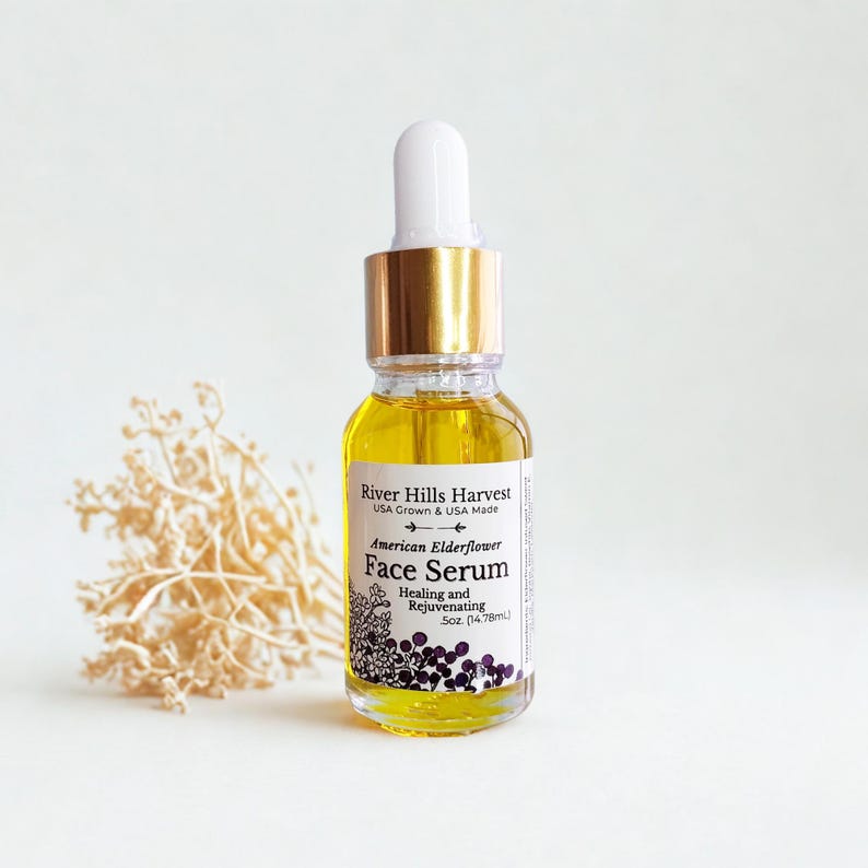 May include: A clear glass bottle of American Elderflower Face Serum with a white dropper and gold cap. The label reads "River Hills Harvest" and "Healing and Rejuvenating". The bottle contains a yellow liquid and is 0.5 oz (14.78ml).