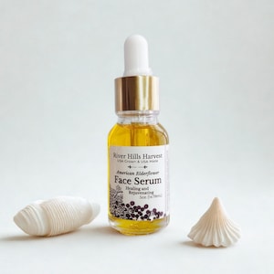 May include: A clear glass bottle of face serum with a gold cap and white dropper. The label reads "River Hills Harvest American Elderflower Face Serum." Two white seashell decorations are on either side of the bottle. The serum is 0.5 fl oz (14.78ml).
