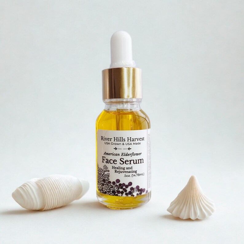 May include: A clear glass bottle of face serum with a gold cap and white dropper. The label reads "River Hills Harvest American Elderflower Face Serum." Two white seashell decorations are on either side of the bottle. The serum is 0.5 fl oz (14.78ml).