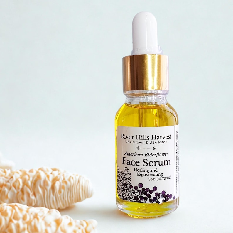 May include: A clear glass bottle of face serum with a white dropper and gold cap. The label reads "River Hills Harvest American Elderflower Face Serum." The serum is a yellow liquid. The bottle contains 14.78ml of product.