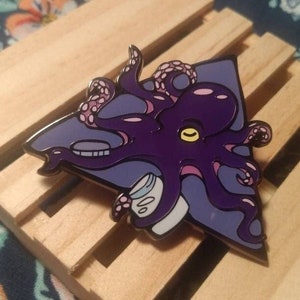 Op de afbeelding: Paarse octopus emaille pin met een roze en blauwe achtergrond. De octopus houdt een klein pilletjesflesje vast. De pin heeft een driehoekige vorm.
