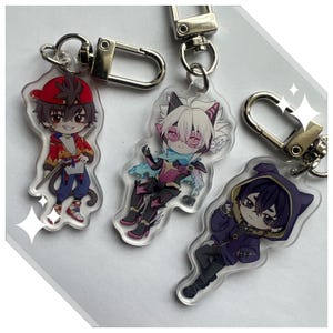 May include: Three anime-style acrylic keychains featuring colorful character designs. Each keychain has a silver-colored clasp. The characters are depicted in various outfits, with one wearing a red cap and jacket.