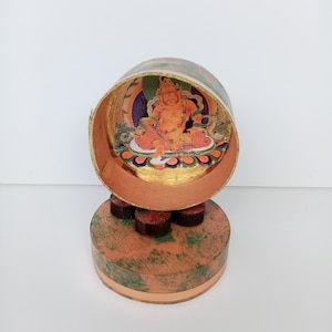 Könnte beinhalten: Eine kleine, runde, goldbemalte Schachtel mit einem bunten Bild einer Gottheit im Inneren. Die Schachtel steht auf einer kleineren, runden, braunen Schachtel mit grünen Akzenten.