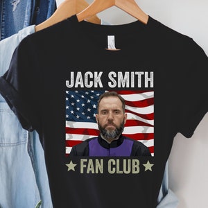 Op de afbeelding: Zwart T-shirt met een witte en rode Amerikaanse vlag afbeelding en de tekst "JACK SMITH FAN CLUB" in wit. Het gezicht van een man is in het midden van de vlag.