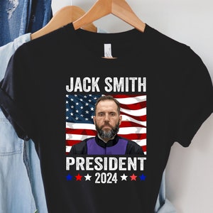 Op de afbeelding: Zwart T-shirt met een afbeelding van Jack Smith, een man met een baard, voor een Amerikaanse vlag. De tekst "Jack Smith President 2024" staat onder de afbeelding.