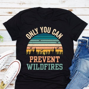 以下が含まれることがあります： 「Only You Can Prevent Wildfires」というテキストと、ヴィンテージスタイルの夕焼けのグラフィックがプリントされた黒いTシャツです。