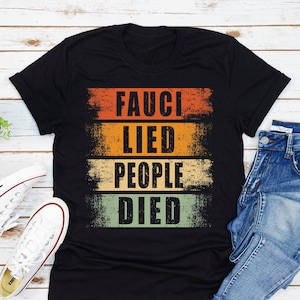 May include: Black t-shirt with the text "FAUCI LIED PEOPLE DIED" in a distressed, retro style. The text is layered in orange, yellow, and green horizontal bands. The shirt is paired with blue jeans and white sneakers.