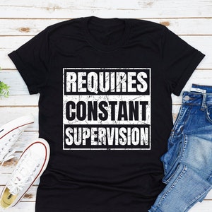 May include: Black t-shirt with white text that reads "REQUIRES CONSTANT SUPERVISION" in a distressed font.