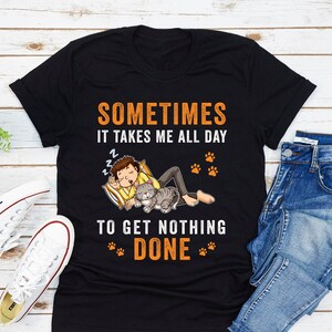 May include: Black t-shirt with a humorous saying about getting nothing done. The text reads "Sometimes it takes me all day to get nothing done."  The graphic features a person sleeping with a cat.