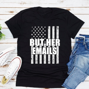 May include: Black t-shirt with a distressed American flag design and the text "BUT HER EMAILS" in white.