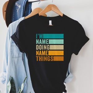 May include: Black t-shirt with a retro-style graphic that reads "I'm NAME doing NAME things". The graphic is made up of colorful stripes in shades of teal, green, beige, and orange.