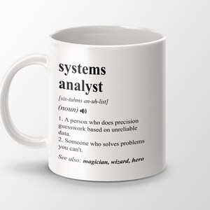 Puede incluir: Taza de cerámica blanca con un diseño negro que define a un analista de sistemas como una persona que hace conjeturas precisas basadas en datos poco fiables y alguien que resuelve problemas que no puedes resolver. El texto también incluye la pronunciación fonética de la palabra "systems analyst" y las palabras "Ver también: mago, mago, héroe".