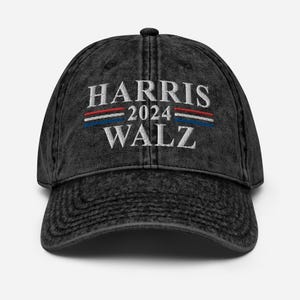 Puede incluir: Gorra de béisbol negra de aspecto desgastado con el texto bordado "HARRIS 2024 WALZ" en blanco. Encima del texto, rayas horizontales rojas, blancas y azules. La gorra tiene una visera curva y un botón en la parte superior.