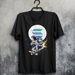May include: Black t-shirt with a graphic of an astronaut riding a skateboard in front of a moon with a blue and green logo. The astronaut is wearing a white helmet and holding a cup.