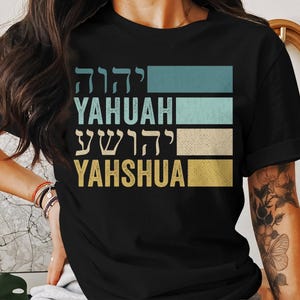 May include: Black t-shirt with a vintage-style design featuring the Hebrew words for "Yahweh" and "Yahshua" in a colorful striped pattern.