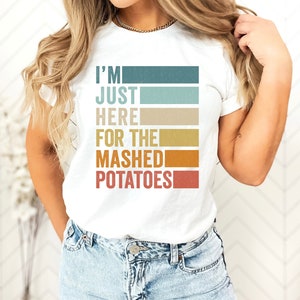 Può includere: Maglietta bianca con una grafica in stile retrò che dice "I'm Just Here For The Mashed Potatoes".
