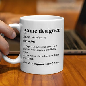 Puede incluir: Taza de cerámica blanca con un diseño negro que dice "game designer" y define el término como "una persona que hace conjeturas precisas basadas en datos poco fiables". La definición también incluye "Ver también: mago, mago, héroe."