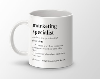 May include: White ceramic mug with a black and white definition of "marketing specialist" printed on it. The definition includes the phonetic pronunciation, a definition of the word, and a list of related words: "magician, wizard, hero".