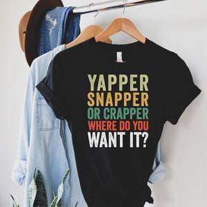 May include: Black t-shirt with the text "YAPPER SNAPPER OR CRAPPER WHERE DO YOU WANT IT?" in a retro font. The shirt is hanging on a wooden hanger with a light blue denim shirt and a brown hat in the background.
