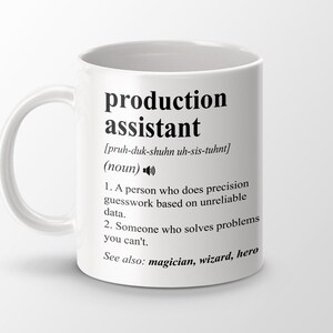 May include: White ceramic mug with a black design that defines the word "production assistant" and its pronunciation. The definition includes two bullet points and the phrase "See also: magician, wizard, hero".