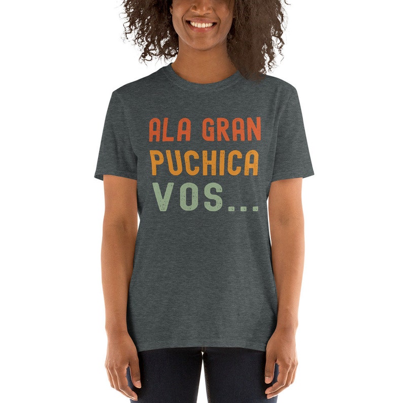 May include: A dark heather grey t-shirt with the text "ALA GRAN PUCHICA VOS..." printed in a retro style with orange, yellow, and green colors.