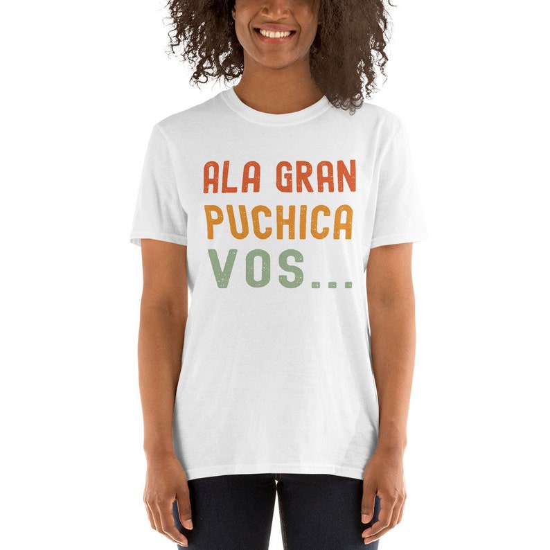 May include: White t-shirt with a vintage-style graphic that reads "ALA GRAN PUCHICA VOS..." in red, orange, and green.